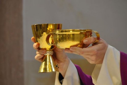 Ein goldener Kelch und eine goldene Hostienschale werden von zwei Armen im Messgewand hochgehalten.