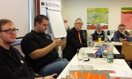 Fünf Männer sitzen rund um einen Tisch. Im Hintergrund sieht man das Logo des Kolpingverbandes und ein Flipchart.