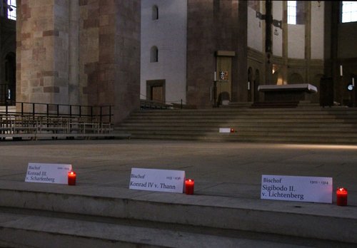 Rote Kerzen stehen auf dem Boden im Königschor des Speyerer Domes vor Namensschildern von im Dom bestatteten Bischöfen